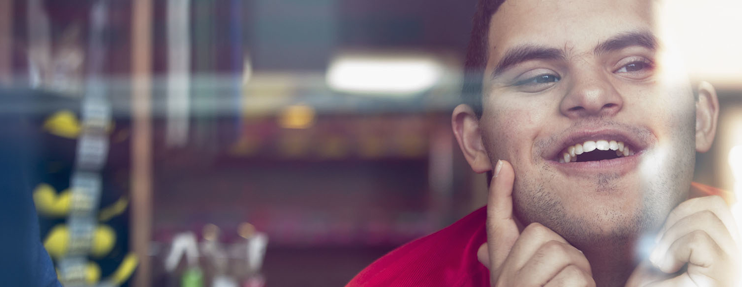 A young man with disabilities looks out through a window, smiling, with both his hands on his face